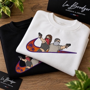 May include: A white and a black crewneck sweatshirt, each featuring an embroidered graphic of two figures wearing bandanas and holding handguns, positioned inside a large orange and purple swoosh logo decorated with palm tree silhouettes.