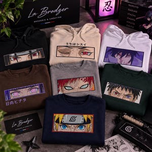 Può includere: Una collezione di sette felpe con e senza cappuccio in nero, beige, bianco, marrone, grigio, verde scuro e blu navy. Ogni capo presenta un grafico rettangolare ricamato con occhi di personaggi anime e testo giapponese sopra o sotto il disegno.