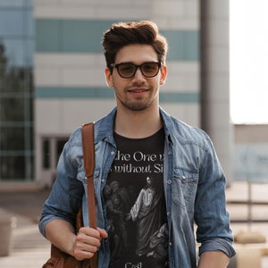 May include: A man wearing a black t-shirt with a graphic of Jesus and the text "The One without Sin". He is wearing a blue denim jacket and brown leather shoulder bag.