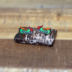 May include: Two green, red, and blue painted earrings shaped like a stylized dragon head with sharp teeth. The earrings are on a dark brown, textured surface.