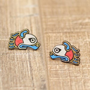 May include: A pair of wooden stud earrings featuring a stylized fish design. The fish have a white face, red accents, and blue and gold details. The earrings are displayed on a light brown wooden surface.