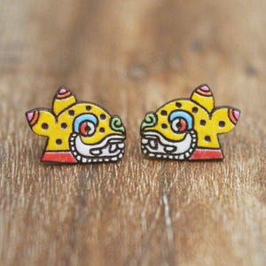 May include: A pair of yellow wooden earrings with a colorful design of a jaguar face. The earrings are hand-painted with details in red, blue, green, and white.