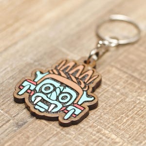 May include: Wooden keychain featuring a stylized, colorful face design. The face has a light blue face, brown hair, and red accents. The keychain has a silver metal ring and chain.