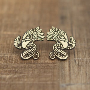 May include: A pair of wooden earrings featuring a detailed design of a mythical creature with a feathered headdress and a serpent-like body. The earrings are painted in a light cream color with black outlines.