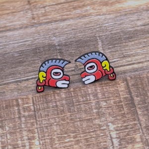 May include: Two small, colorful pins featuring a stylized face design. The faces are red with white, yellow, and gray accents, and black outlines. The pins are displayed on a wooden surface.