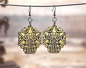 May include: A pair of gold-colored, star-shaped earrings with a black outline. Each earring features a stylized face design with a headdress and a patterned lower section. The earrings hang from black metal hooks.