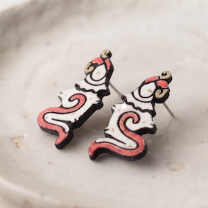 May include: A pair of stud earrings featuring a stylized, abstract creature design. The earrings are crafted with a black outline, white interior, and vibrant red accents, showcasing a curved, serpentine shape with a small circular head detail.