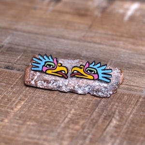 May include: A pair of colorful wooden earrings featuring a stylized bird design with blue, pink, yellow, and green accents. The earrings are displayed on a light brown stone surface.