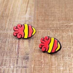 May include: Two pairs of red, yellow, and black earrings shaped like a squid or octopus. The earrings have a red, swirled top and a yellow and black striped body. They are set on a wooden surface.
