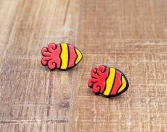May include: Two pairs of red, yellow, and black earrings shaped like a squid or octopus. The earrings have a red, swirled top and a yellow and black striped body. They are set on a wooden surface.