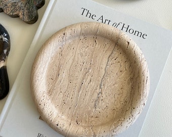 May include: A beige travertine stone tray with a round shape and a slightly raised rim. The tray is sitting on a white book with the text "The Art of Home" and "Written by Shea McGee".