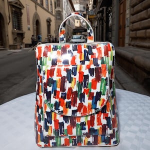 Puede incluir: Una mochila blanca con un diseño abstracto de salpicaduras de pintura de colores. La mochila tiene un bolsillo frontal grande y un cierre de cremallera.