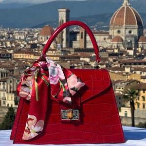 May include: A red crocodile embossed leather handbag with a floral patterned silk scarf. The handbag has a top handle and a gold clasp.