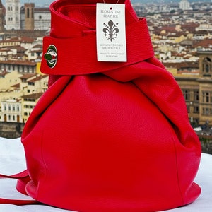 Peut inclure: Un sac à dos en cuir rouge vif avec une fermeture à cordon et une boucle argentée. Le sac à dos porte une étiquette indiquant "Florentine Leather, Genuine Leather Made in Italy, Prodotto Artigianale Fiorentino".