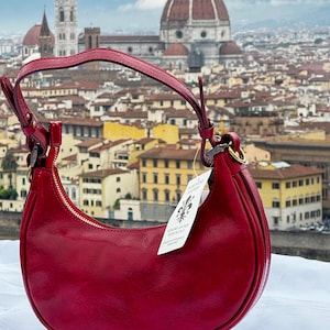 May include: A red leather handbag with a gold zipper and a gold tag that says "FIRENZE LEATHER MADE IN ITALY". The bag is in front of a cityscape with a large dome building in the background.