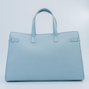 May include: A light blue handbag with two top handles and a structured design. The bag features a textured surface and a rectangular shape with a flap closure on each side. The bag has a light blue color.