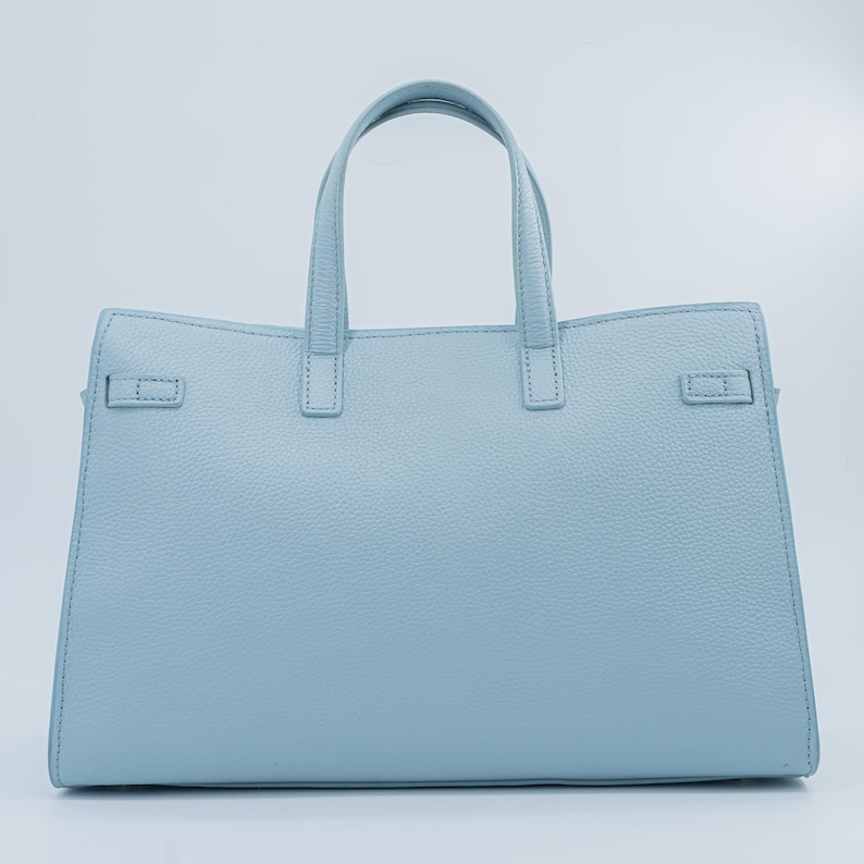May include: A light blue handbag with two top handles and a structured design. The bag features a textured surface and a rectangular shape with a flap closure on each side. The bag has a light blue color.
