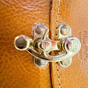 May include: Close-up of a gold-toned metal clasp with a decorative design. The clasp is attached to a brown leather surface with stitching.