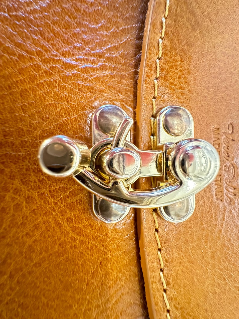 May include: Close-up of a gold-toned metal clasp with a decorative design. The clasp is attached to a brown leather surface with stitching.
