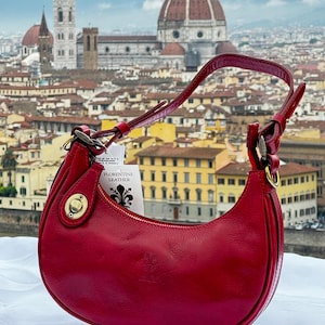May include: A red leather handbag with a gold clasp and a tag that reads "Florentine Leather". The bag is photographed in front of a cityscape with a large dome building in the background.