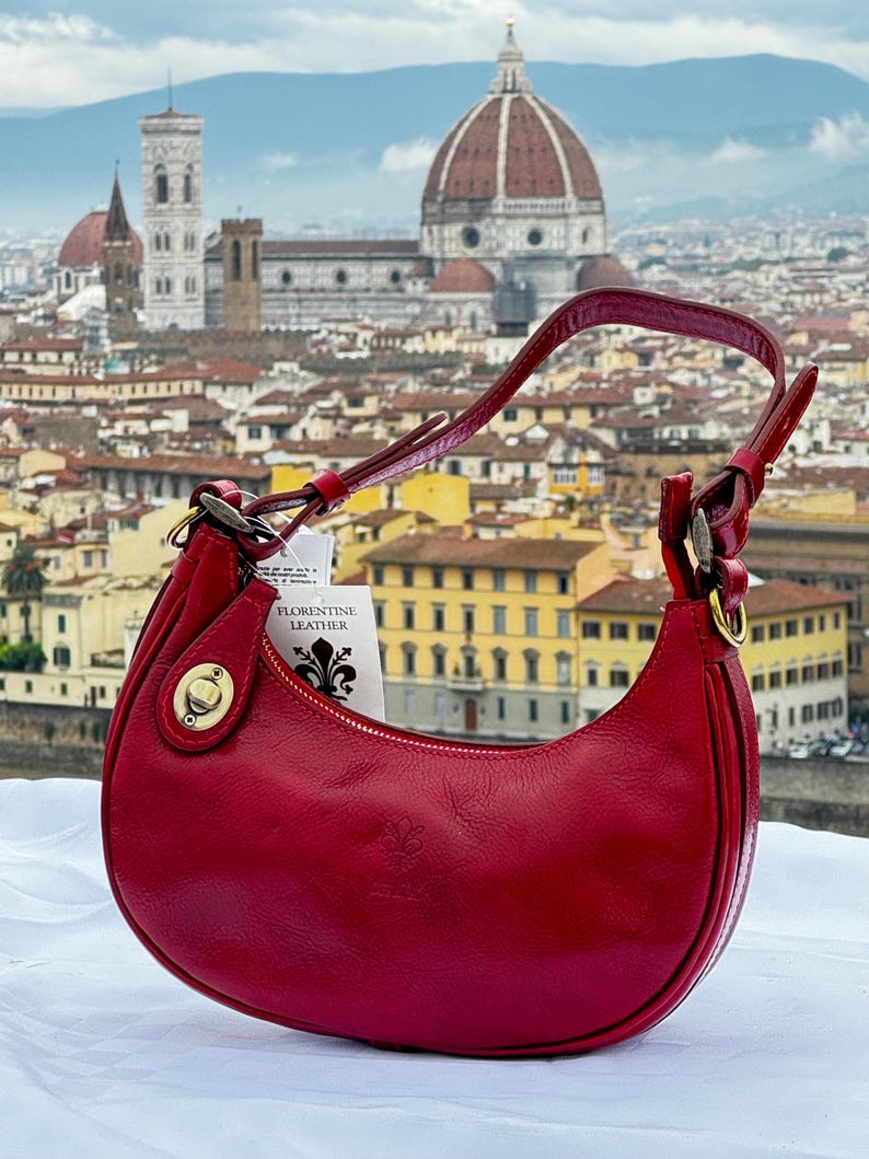 May include: A red leather handbag with a gold clasp and a tag that reads "Florentine Leather". The bag is photographed in front of a cityscape with a large dome building in the background.