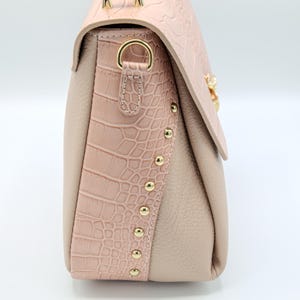 May include: A blush pink handbag with a textured crocodile-style flap and side panel. The bag features gold-colored studs and hardware, with a soft, neutral-toned body. The bag has a structured design and a clasp closure.