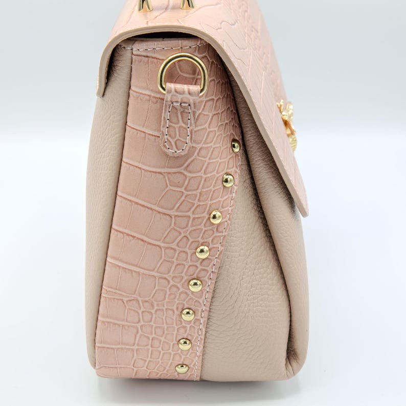 May include: A blush pink handbag with a textured crocodile-style flap and side panel. The bag features gold-colored studs and hardware, with a soft, neutral-toned body. The bag has a structured design and a clasp closure.