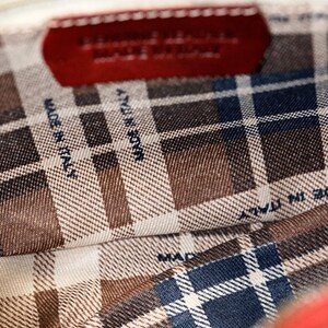 May include: The inside of a red leather bag with a plaid lining. The lining has the text "Made in Italy" printed on it.