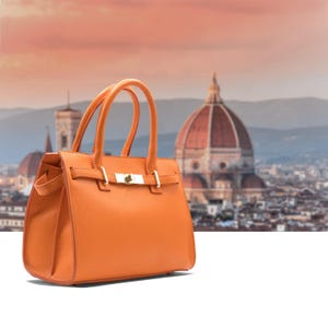 May include: Orange leather handbag with a top handle and gold-tone hardware. The bag features a structured design with a clasp closure. The background shows a blurred cityscape with a prominent dome.