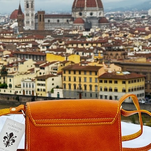 May include: A small, orange leather crossbody bag with a yellow strap. The bag has a flap closure and a small pocket on the front. The bag is in front of a blurry background of the city of Florence, Italy. The tag on the bag reads "Florentine Leather" and "Genuine Leather Made in Italy."