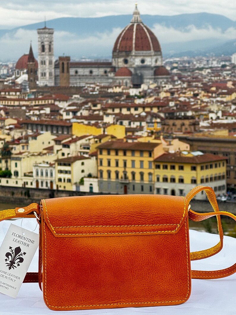 May include: A small, orange leather crossbody bag with a yellow strap. The bag has a flap closure and a small pocket on the front. The bag is in front of a blurry background of the city of Florence, Italy. The tag on the bag reads "Florentine Leather" and "Genuine Leather Made in Italy."
