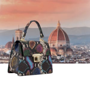 Op de afbeelding: Een veelkleurige handtas met slangenpatroon, een handvat aan de bovenkant en goudkleurige hardware. De tas heeft een rechthoekige vorm met een klepsluiting. De achtergrond toont een vage stadsgezicht met een groot koepelvormig gebouw.