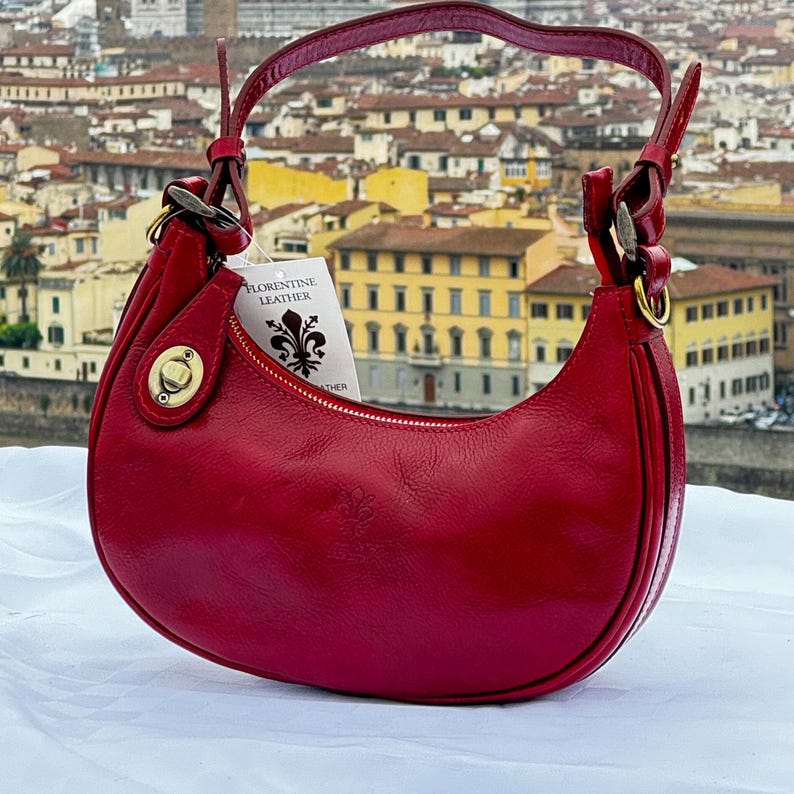 A sleek red leather handbag is elegantly displayed on a table, with the historic architecture of Florence, including the iconic Duomo, visible in the background. The bag's shiny finish and craftsmanship reflect the luxury of Italian design