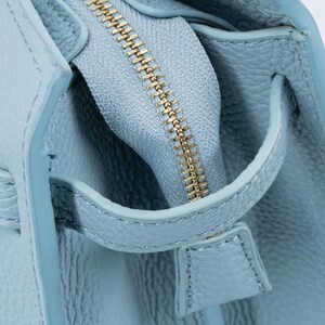 May include: Close-up of a light blue leather handbag with a gold zipper. The zipper is partially open, revealing the interior. The leather has a textured grain, and the handle is visible.
