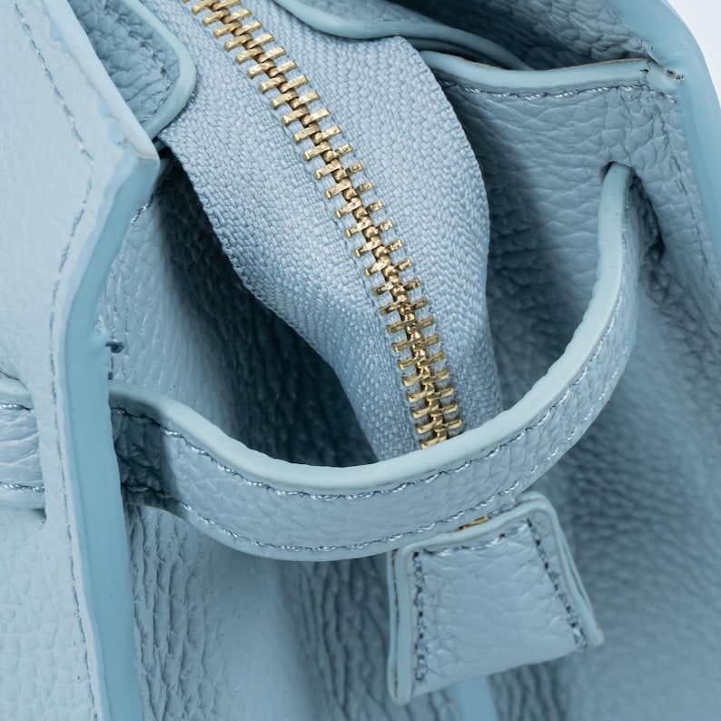 May include: Close-up of a light blue leather handbag with a gold zipper. The zipper is partially open, revealing the interior. The leather has a textured grain, and the handle is visible.
