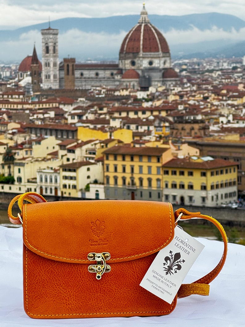 May include: A small, orange leather crossbody bag with a gold clasp. The bag has a tag that reads "Florentine Leather, Genuine Leather Made in Italy, Prodotto Artigianale Fiorentino". The bag is in front of a blurry background of the city of Florence, Italy.