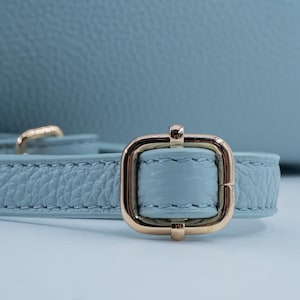 May include: Close-up of a light blue leather strap with a gold-colored rectangular buckle. The strap has a textured surface and is stitched along the edges. The buckle is centered and has a shiny finish.