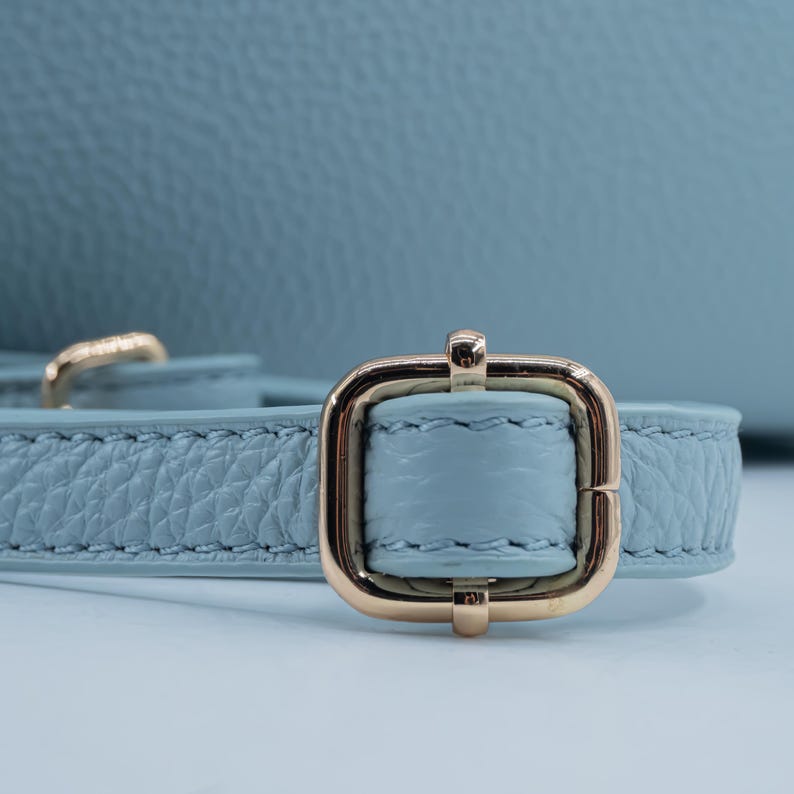 May include: Close-up of a light blue leather strap with a gold-colored rectangular buckle. The strap has a textured surface and is stitched along the edges. The buckle is centered and has a shiny finish.