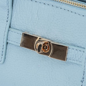 May include: Close-up of a light blue leather handbag with a gold-colored clasp. The clasp is rectangular with a decorative design in the center. The bag has a textured surface and visible stitching.