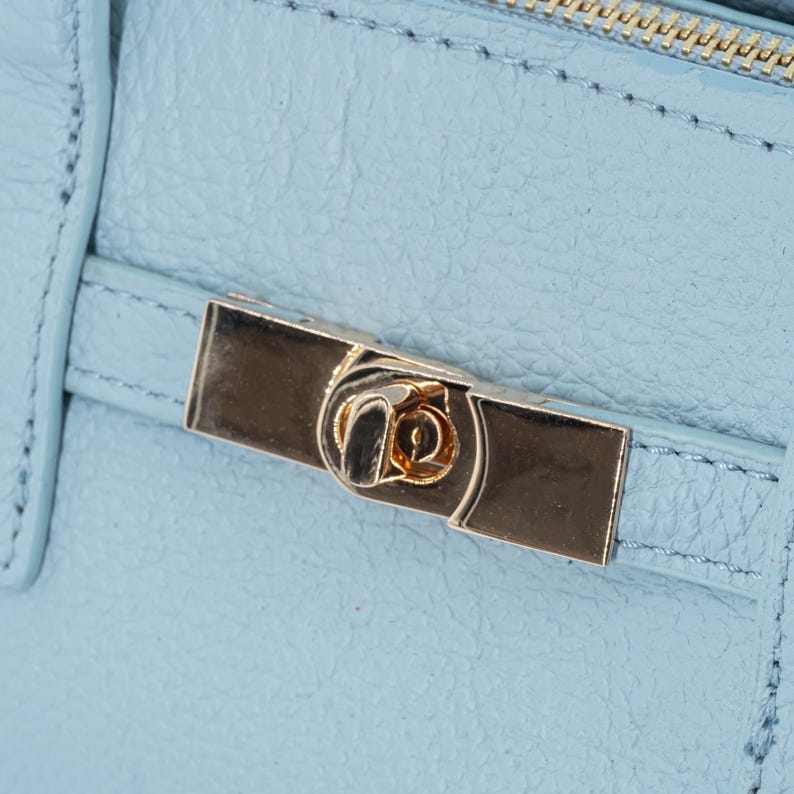 May include: Close-up of a light blue leather handbag with a gold-colored clasp. The clasp is rectangular with a decorative design in the center. The bag has a textured surface and visible stitching.