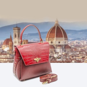 May include: A red and burgundy leather handbag with a top handle and a detachable shoulder strap. The flap has a crocodile-like texture and a gold-coloured bee emblem. Gold-coloured studs adorn the sides of the bag.