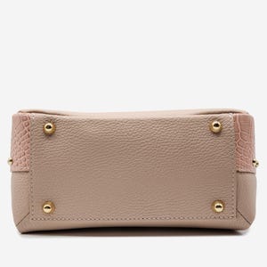 May include: A light pink handbag with a textured leather body and gold-colored metal feet. The corners feature a pink, crocodile-textured leather. The bag has a rectangular shape.