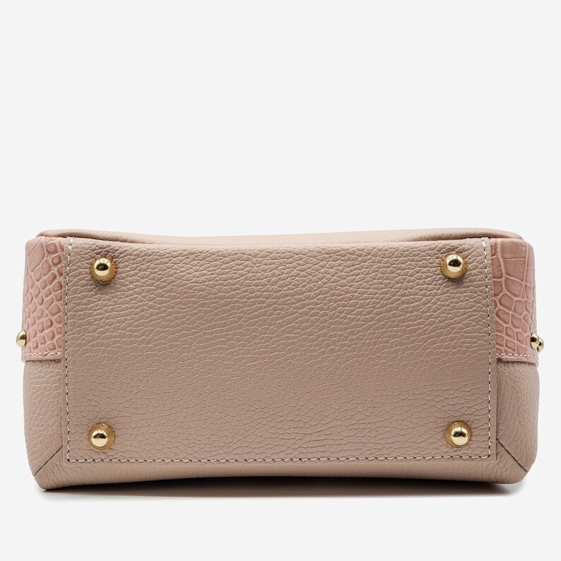 May include: A light pink handbag with a textured leather body and gold-colored metal feet. The corners feature a pink, crocodile-textured leather. The bag has a rectangular shape.