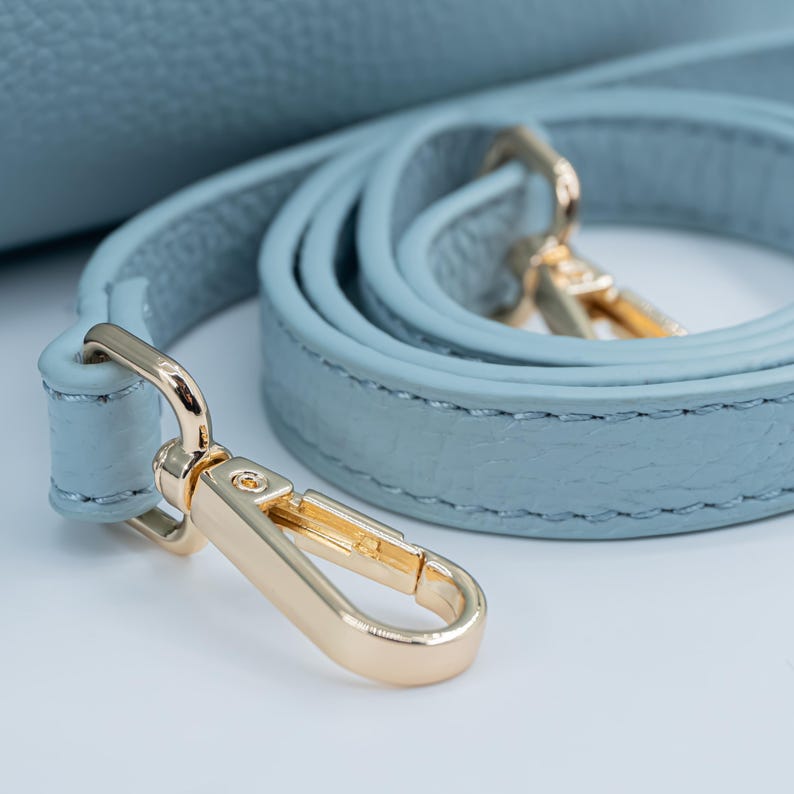 May include: Close-up of a light blue leather handbag strap with gold-colored metal hardware. The strap is coiled, showing the stitching and texture of the leather. The clasp is open, ready to attach to a bag.