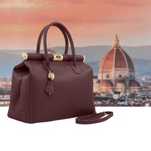 Peut inclure: Un sac à main en cuir bordeaux avec des éléments dorés, dont une serrure et des détails sur les poignées. Le sac a deux poignées supérieures et une bandoulière amovible. En arrière-plan, un paysage urbain flou avec un dôme proéminent.