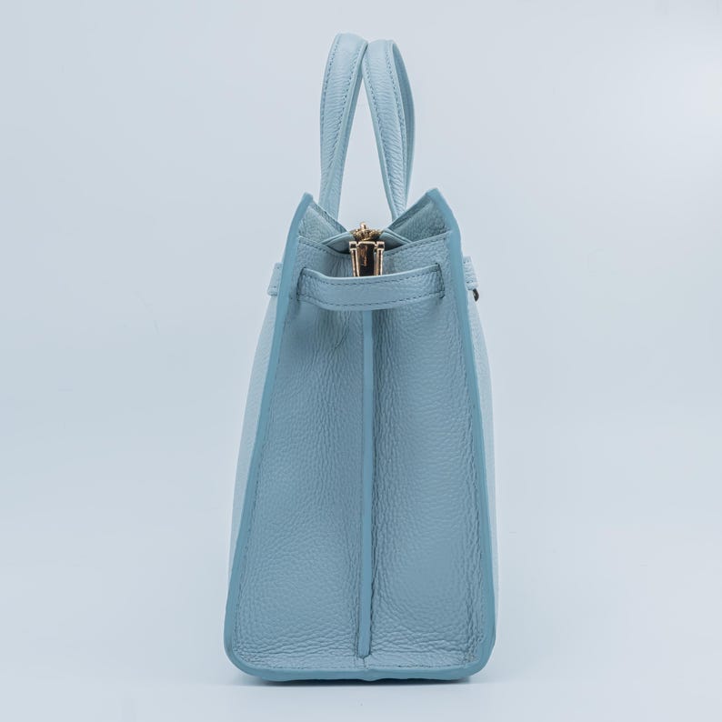 May include: A light blue leather handbag with two handles and a gold zipper. The bag has a structured design with a rectangular base and a slightly tapered shape. The handles are attached to the top of the bag.