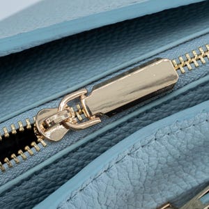 May include: Close-up of a light blue leather item with a gold-colored zipper. The zipper is centered and open, revealing the interior. The leather has a textured appearance, and the zipper pull is rectangular.