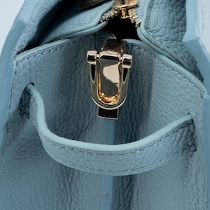 May include: Close-up of a light blue leather handbag with a gold-colored zipper and clasp. The bag has a textured surface and a small handle. The zipper is open, revealing the interior of the bag.