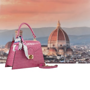 May include: A pink handbag with a crocodile texture, a top handle, and a detachable shoulder strap. The bag features a gold-colored clasp and a decorative scarf tied to the handle. The background shows a cityscape with a large dome.