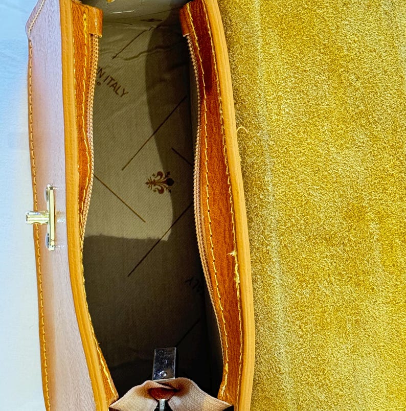 May include: Inside view of a brown leather handbag with a zipper closure. The interior is lined with a beige fabric and has a label that reads "Made in Italy".